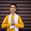 Indian stylish man in yellow traditional clothes with white scarf posed outdoor against wooden background and shows namaste hands sign.